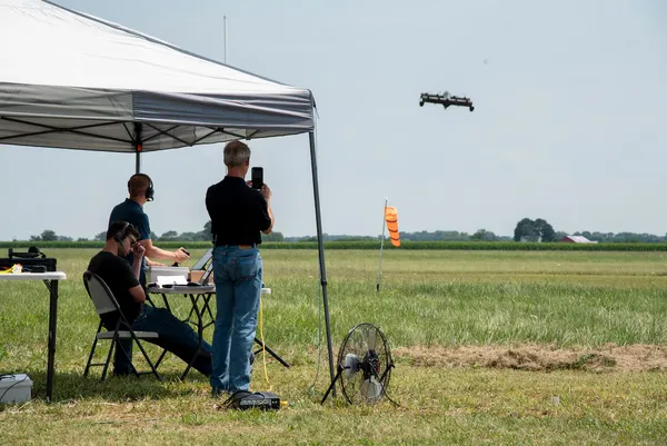 Three Pivotal operators watch from under a tent as they remotely pilot the aircraft over a grassy field.